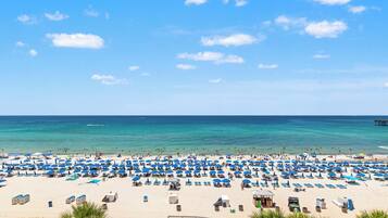 Una playa con numerosas sombrillas azules y sillas de descanso, un muelle de madera a lo lejos y cielos azules despejados.