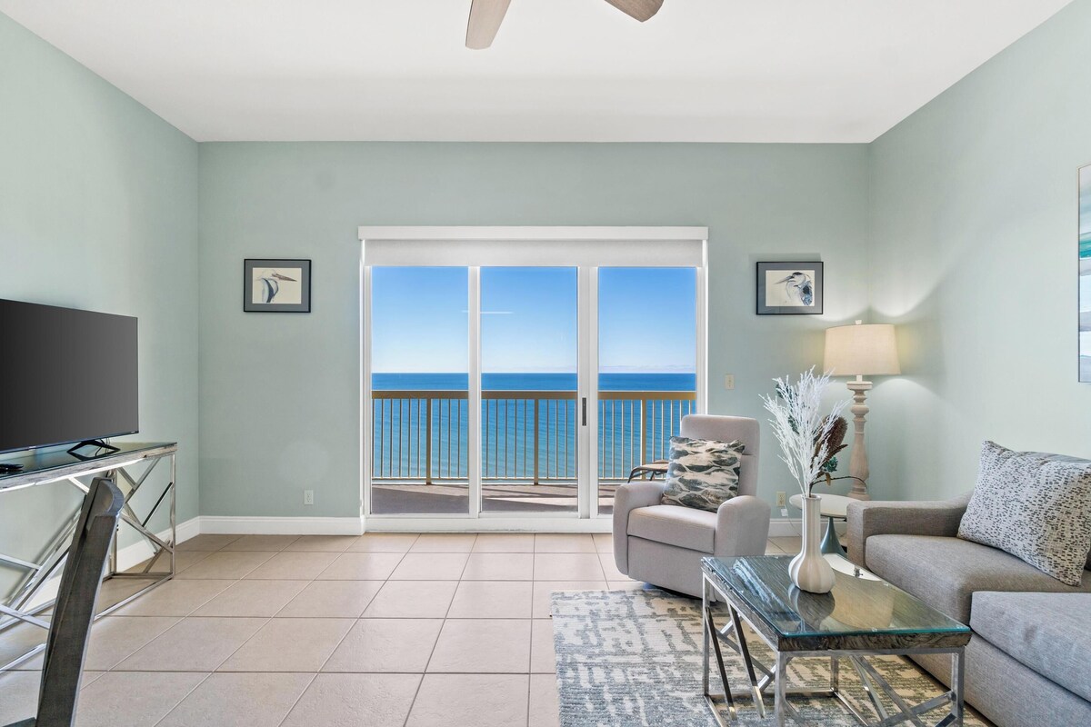 A living room with a television, a sofa, a coffee table, and a view of the ocean through a sliding glass door.