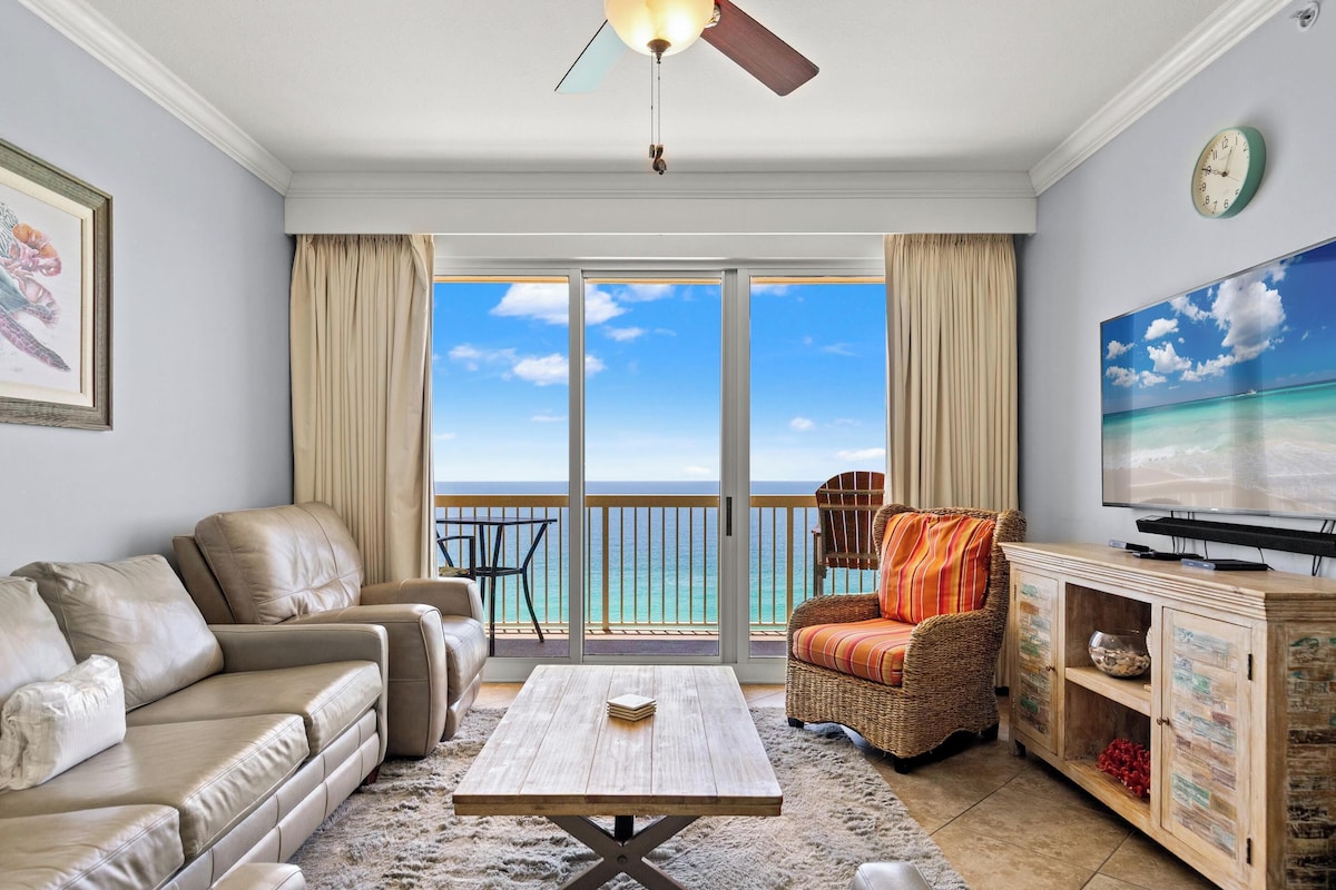 A living room with a sofa, armchair, coffee table, TV, and a view of the ocean through large sliding doors.