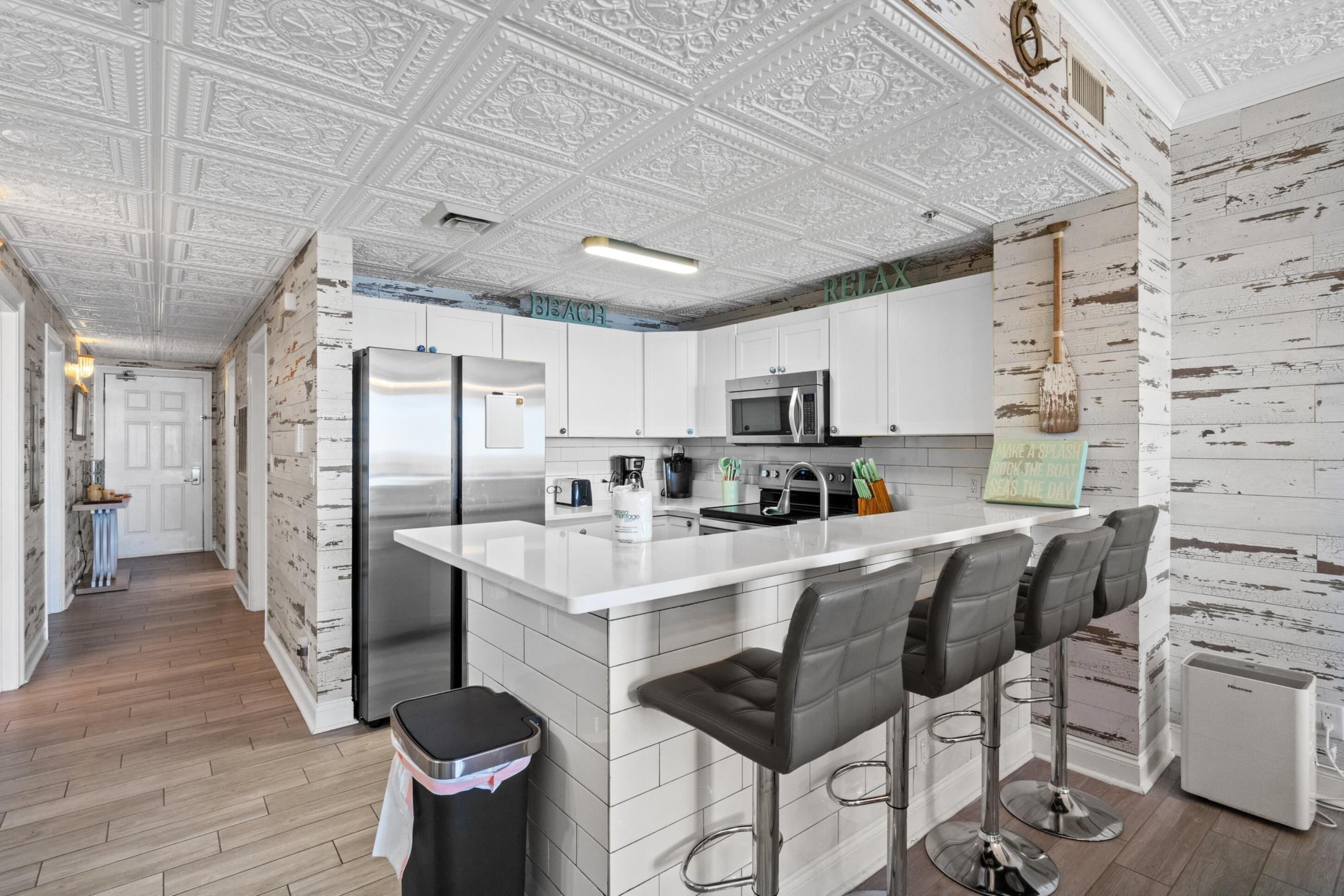 A modern kitchen with white cabinetry, a stainless steel refrigerator, a microwave, and a coffee maker. There are three bar stools and a small island with a sink.