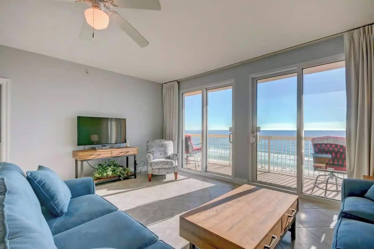 A living room with a sofa, a coffee table, a TV, and a balcony with a view of the sea.