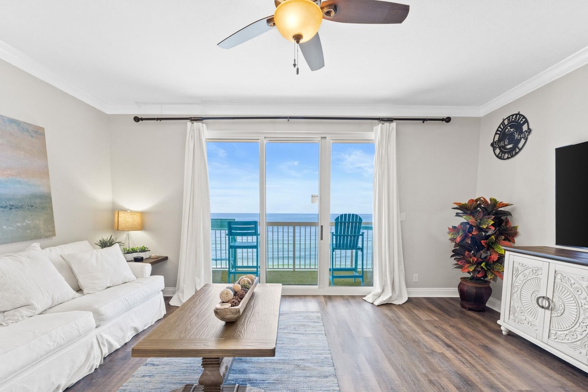 A living room with a sofa, a coffee table, a ceiling fan, a TV, a plant, and a view of the ocean through large sliding doors.