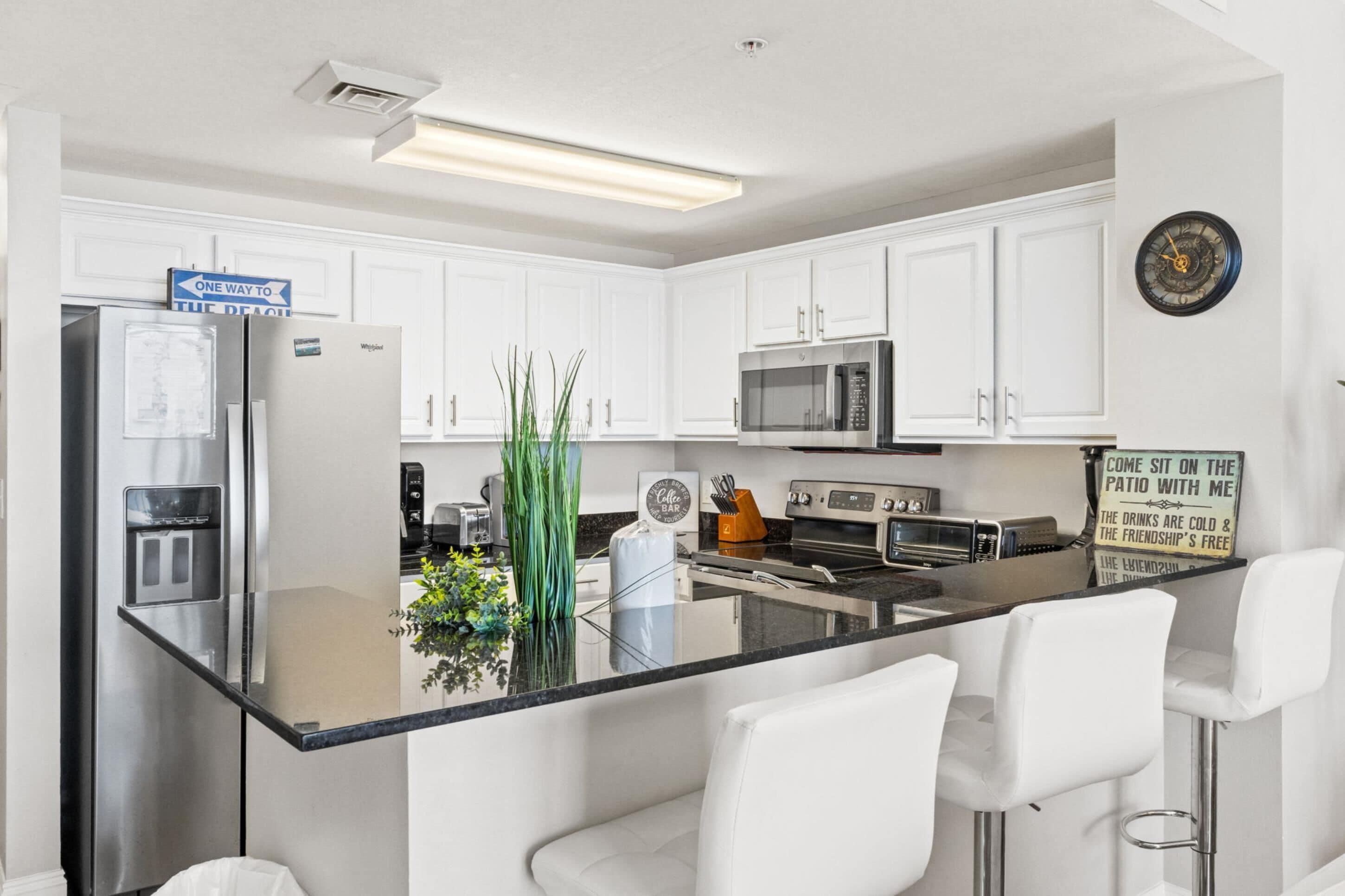 Een moderne keuken met witte kasten, roestvrijstalen apparatuur, een centraal eiland met barkrukken en een klok aan de muur.