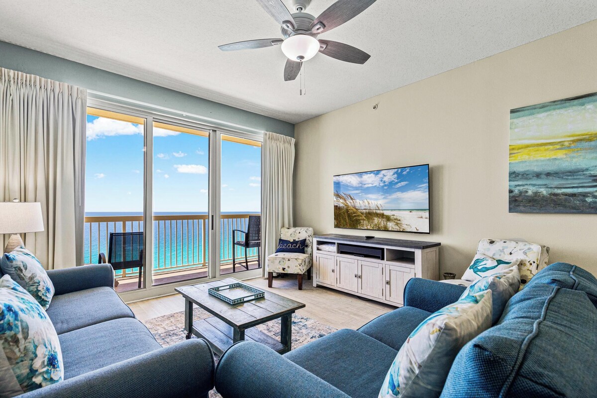 A living room with a sofa, coffee table, TV, and a balcony with a view of the ocean.