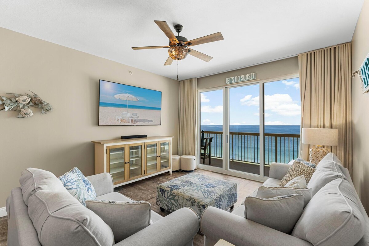 A living room with a television, a ceiling fan, a sofa, a coffee table, and a view of the ocean through large windows.