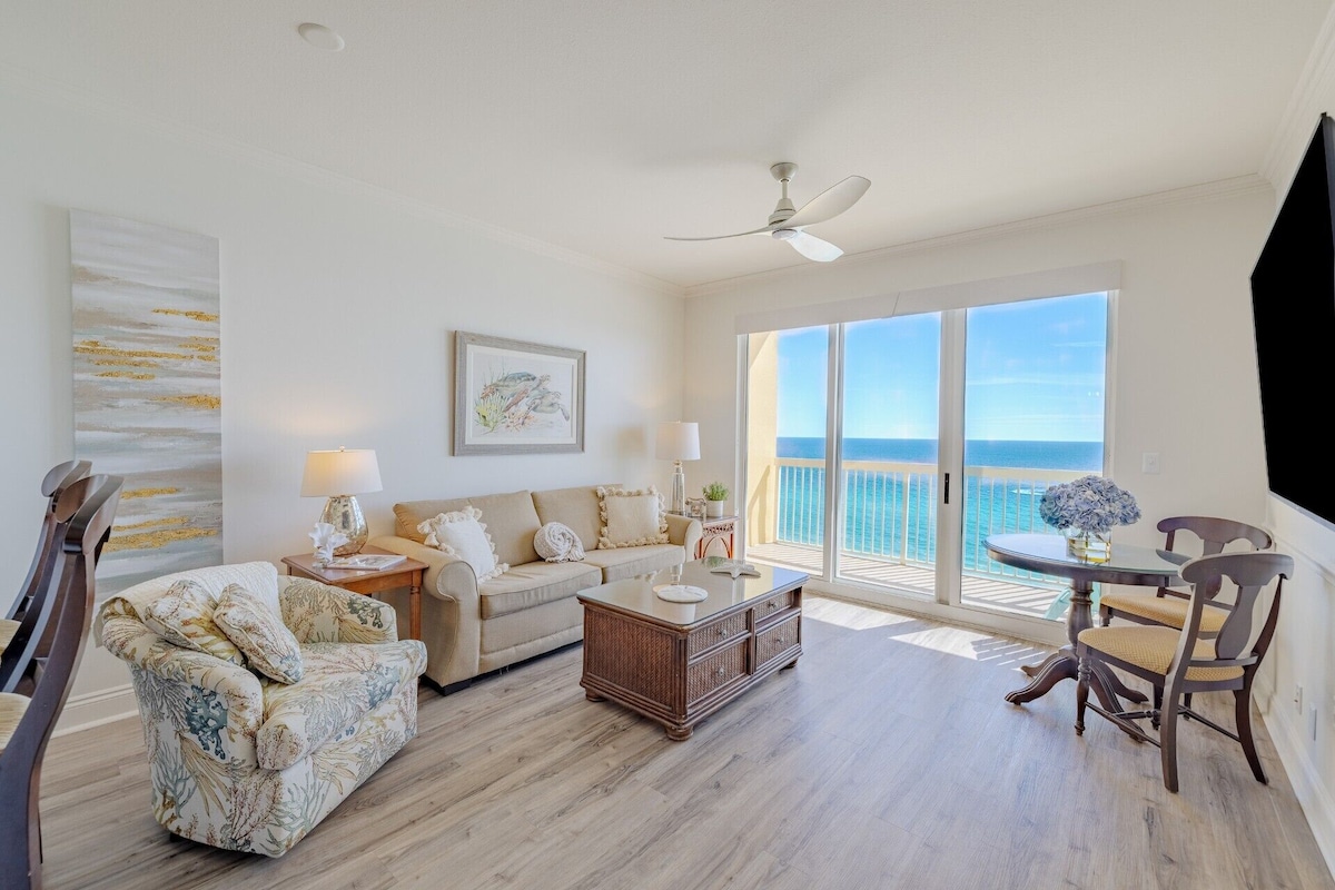 A spacious living room with a balcony overlooking the sea, a ceiling fan, and a large window.
