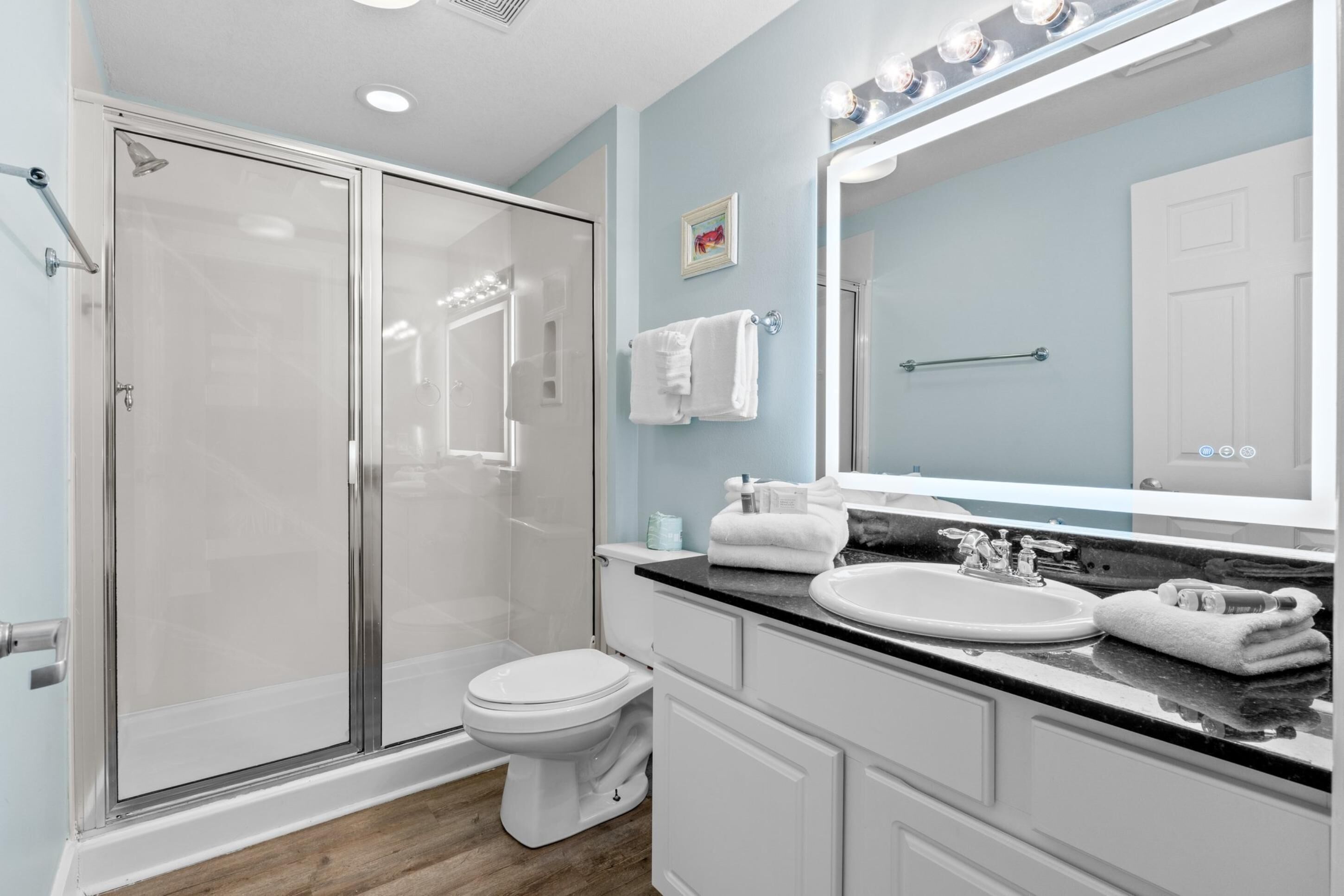 Un baño moderno con ducha de vidrio, inodoro blanco, lavamanos con encimera negra y un espejo grande.