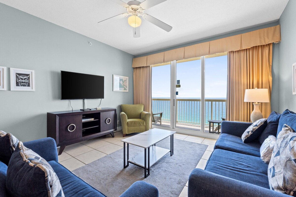 A modern living room with a flat-screen TV, a sofa set, a coffee table, and a view of the ocean through large windows.