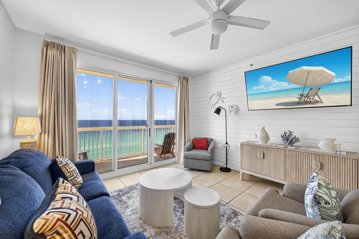 A living room with a sofa, a round coffee table, a TV with a beach scene, a ceiling fan, and a view of the sea through large windows.