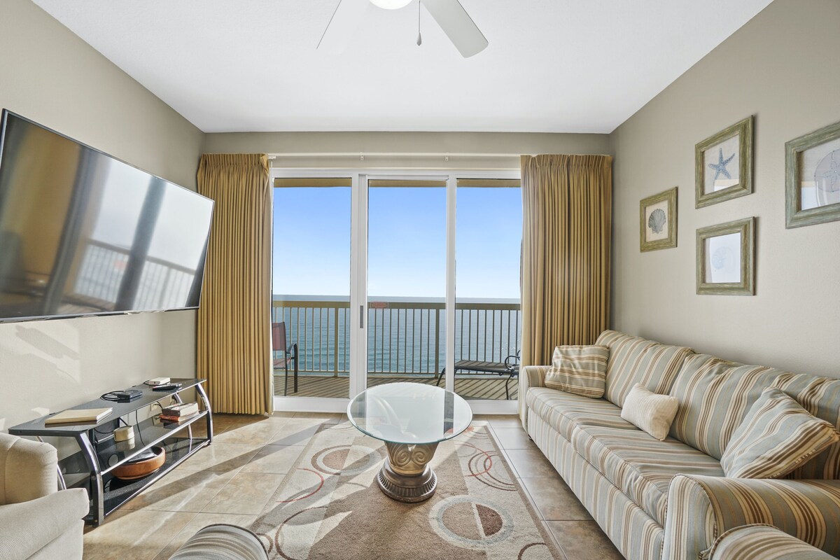 A living room with a sofa, a glass coffee table, a flat-screen TV, and a view of the ocean through large sliding doors.