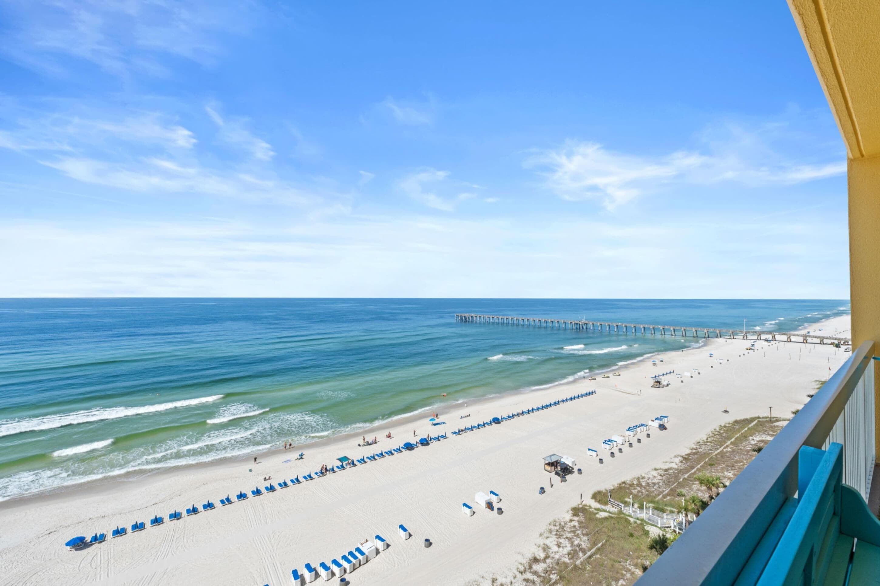 A beach view with a pier extending into the ocean, a sandy beach with lounge chairs, and a clear blue sky.