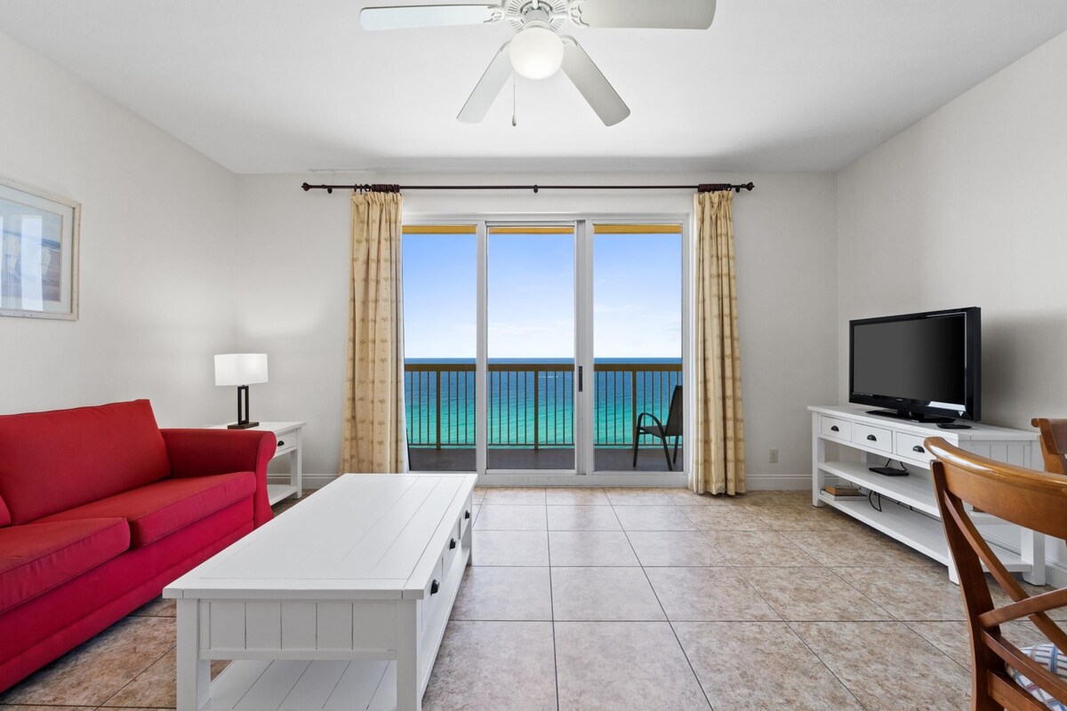 A room with a red sofa, a white coffee table, a TV on a white stand, and a view of the ocean through a sliding glass door.