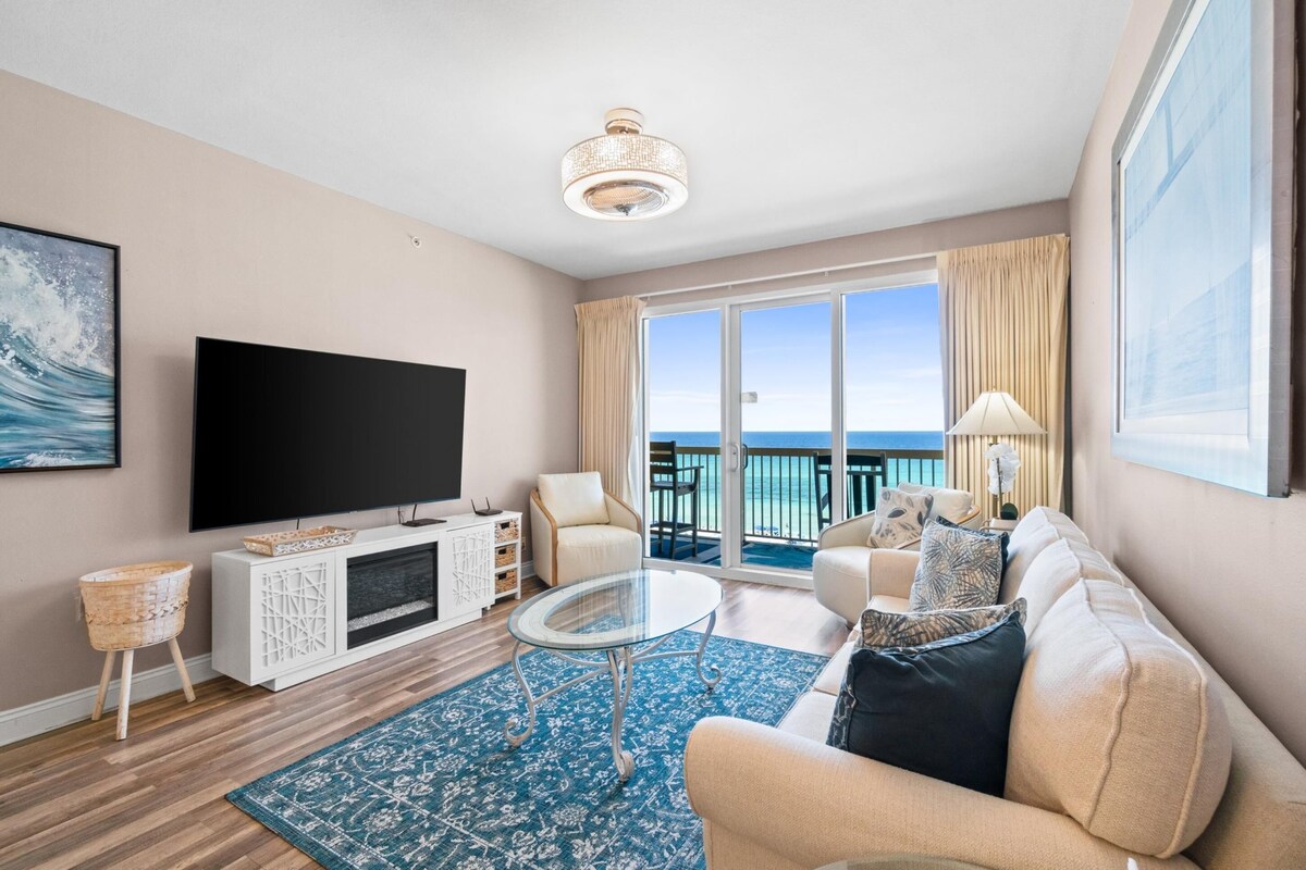 A modern living room with a flat-screen TV, comfortable seating, a glass coffee table, and a view of the ocean through large windows.