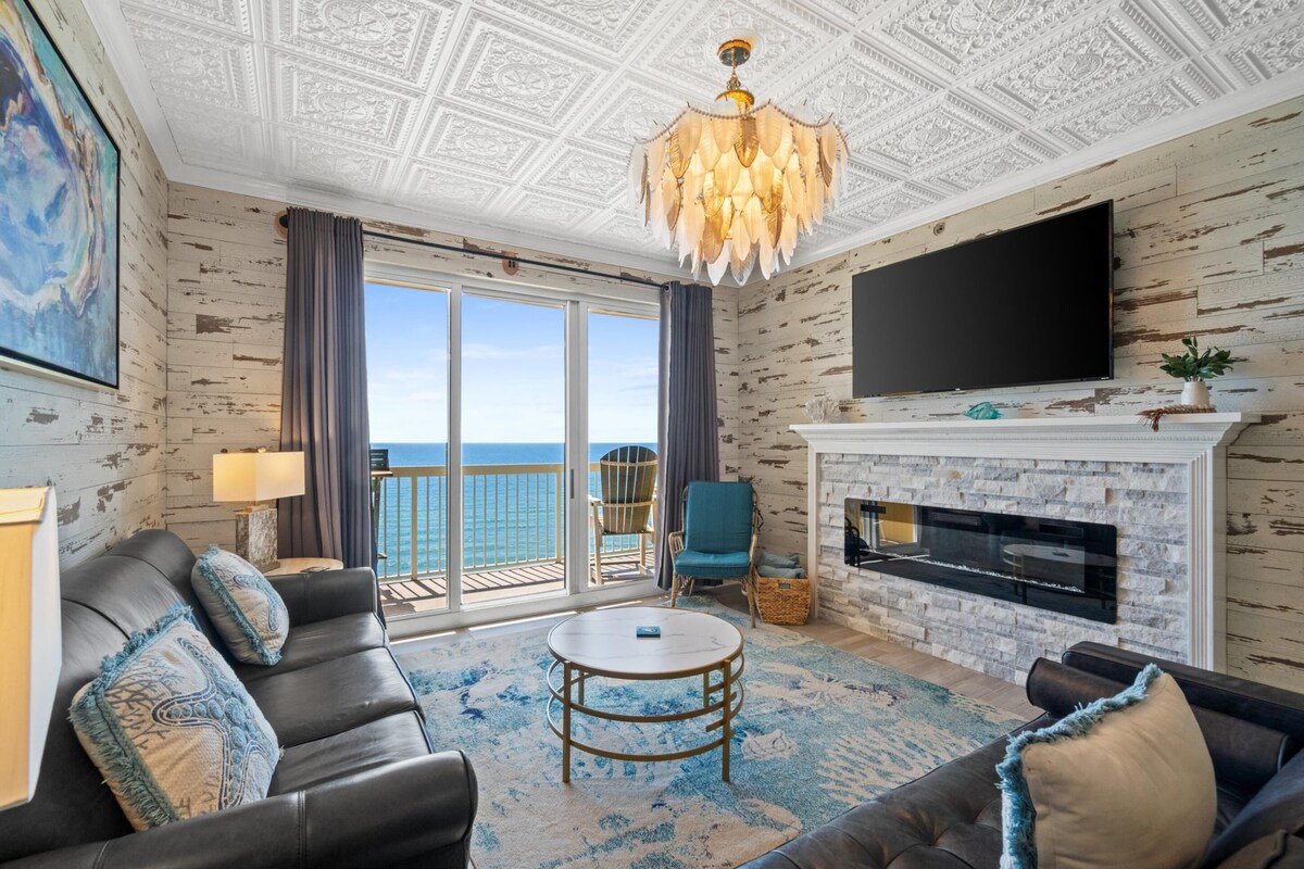 A living room with a fireplace, a large TV, a sofa, and a view of the ocean.