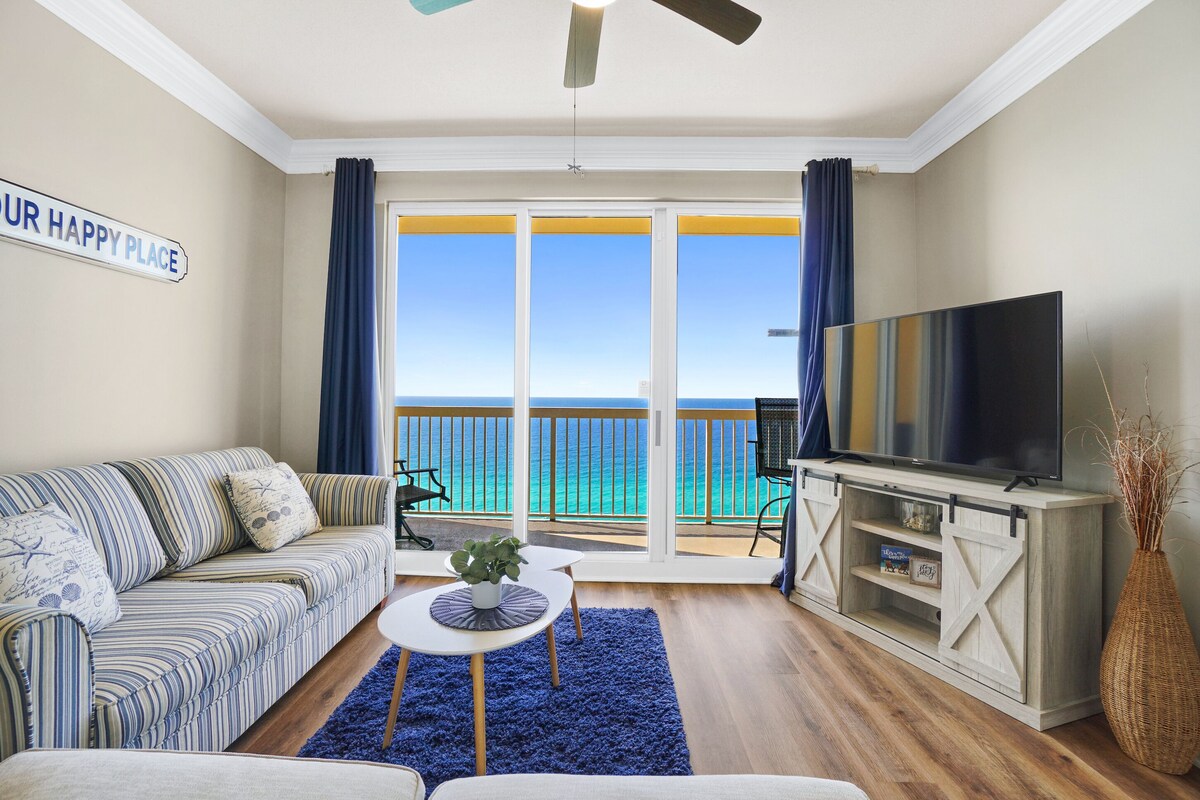 A living room with a sofa, a coffee table, a TV on a wooden cabinet, and a view of the ocean through large sliding doors.