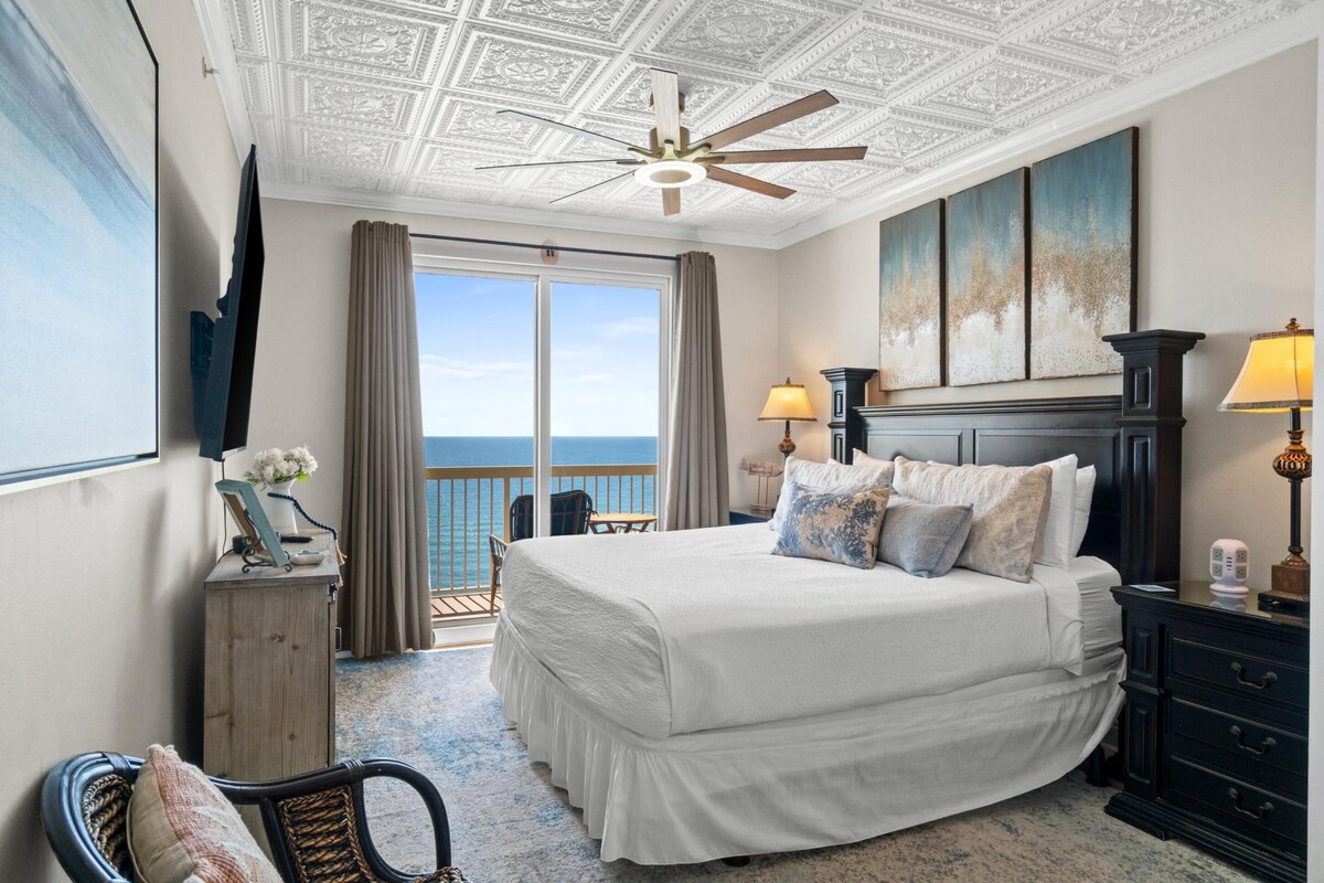 A hotel room with a large bed, a ceiling fan, a TV mounted on the wall, a balcony with a view of the ocean, and a painting above the bed.