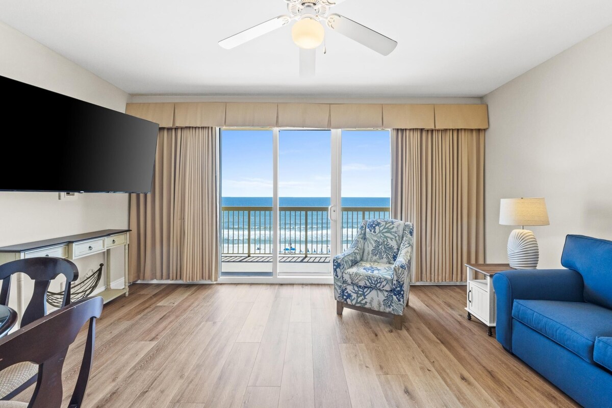 A room with a wooden floor, a blue sofa, a patterned armchair, a TV, a lamp, and a view of the ocean through large windows.