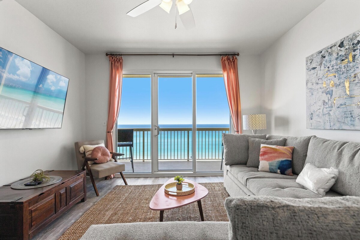 A living room with a sofa, a coffee table, a chair, a TV, and a balcony with a view of the ocean.