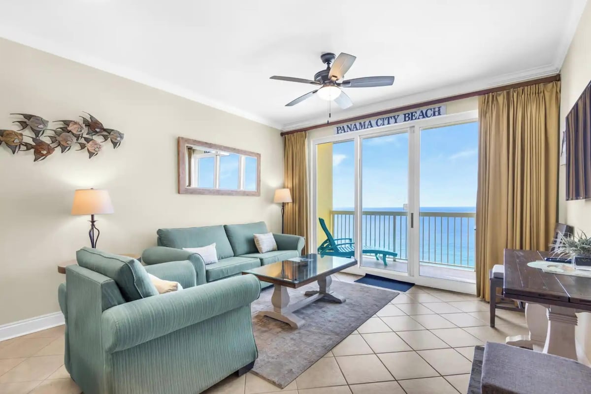 A room with a sofa, a coffee table, a ceiling fan, a view of the ocean, and a sign reading 'Panama City Beach'.