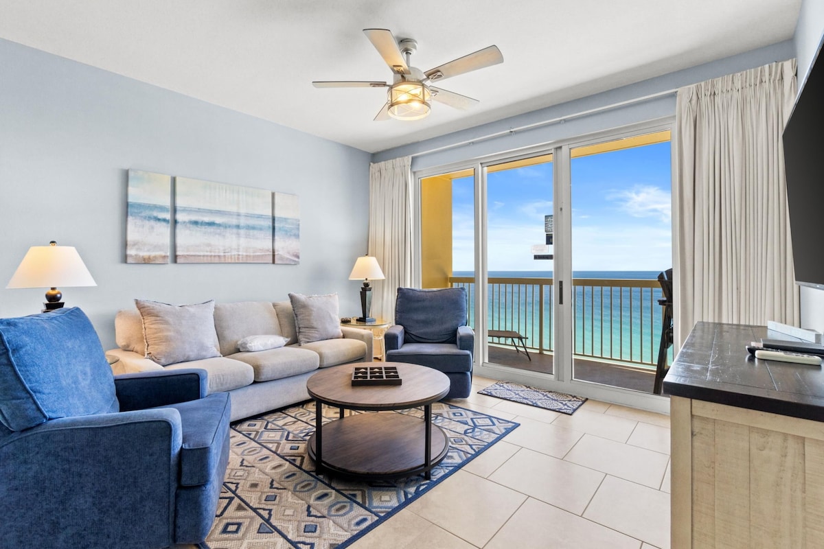 A living room with a sofa, armchairs, a coffee table, a ceiling fan, and a view of the ocean through a sliding glass door.