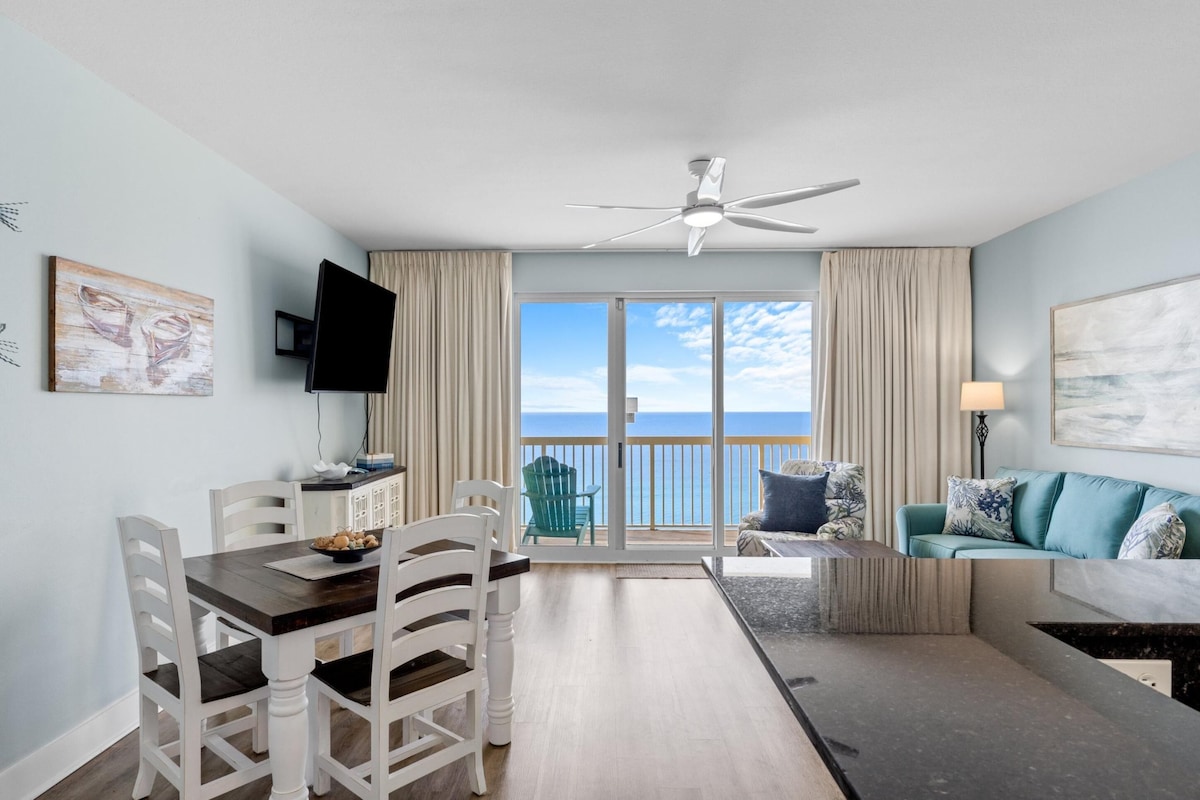 A modern living room with a dining area, a ceiling fan, a TV, and a balcony with a view of the ocean.