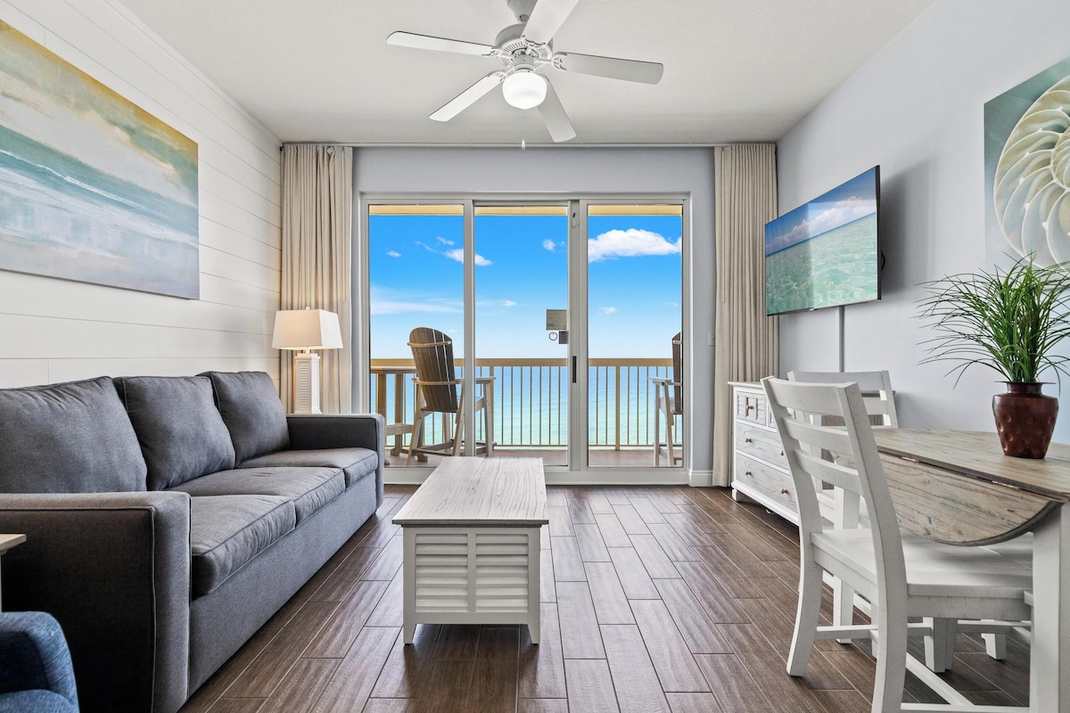 A modern living room with a sofa, a coffee table, a dining table with chairs, a ceiling fan, a TV, and a balcony with a view of the ocean.