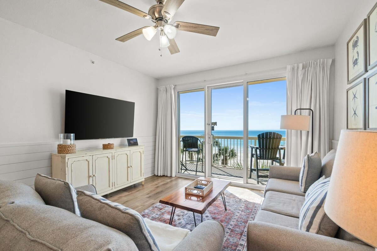 A living room with a flat-screen TV, a ceiling fan, a sofa set, a coffee table, and a view of the ocean through large sliding doors.