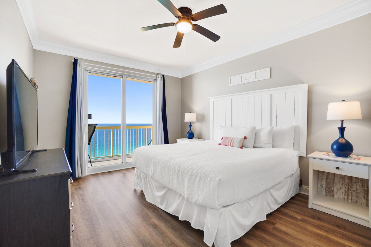 A bedroom with a large bed, a ceiling fan, a TV, and a view of the ocean through a sliding door.