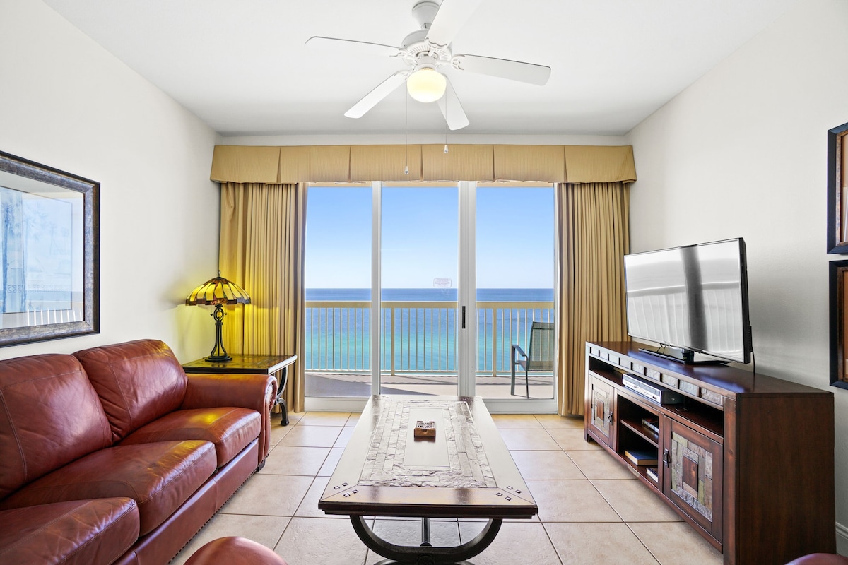 A hotel room with a leather sofa, a TV on a wooden stand, a table with a lamp, and a balcony with a view of the ocean.