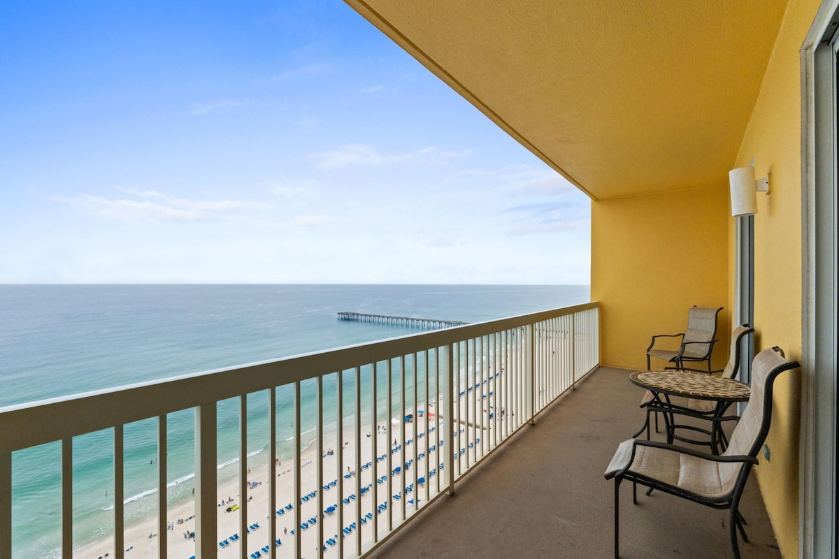 A balcony with a view of the beach and ocean, a table, and two chairs.