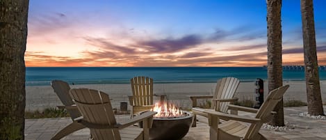 Un patio frente a la playa con sillas Adirondack, un pozo de fuego y palmeras.