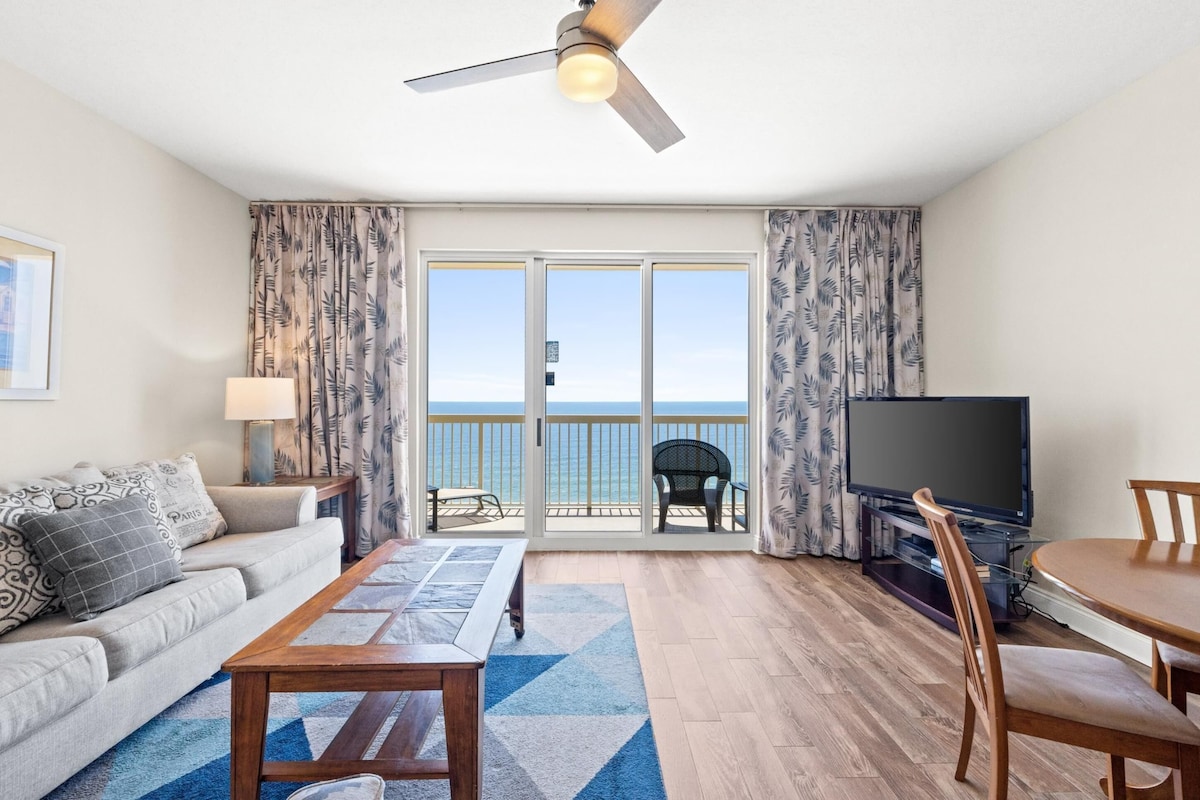 A hotel room with a balcony, a sofa, a TV, a dining table, and a ceiling fan.