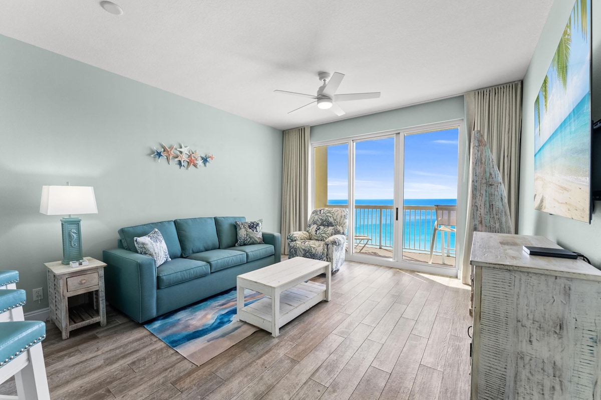 A living room with a teal sofa, a white coffee table, and a view of the ocean through large sliding doors.