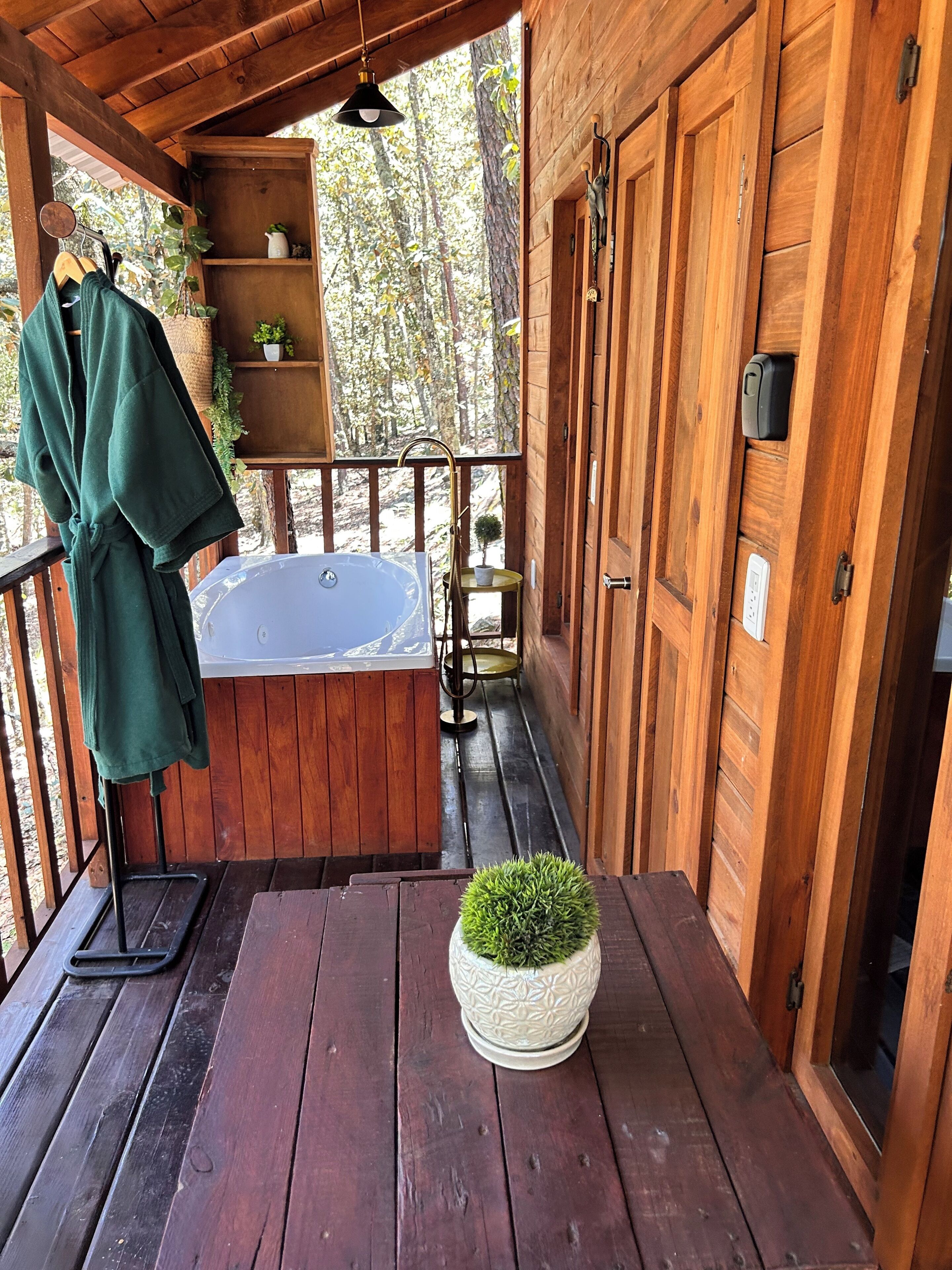 Una cabaña de madera con terraza, jacuzzi, una mesita con planta en maceta y una bata verde colgada en un perchero.