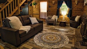 A cozy cabin interior with a wooden staircase, a brown leather sofa, two armchairs, a round rug, and a small table with a lamp. - Peaceful Nature Retreat In Eureka Springs (Eureka Springs)