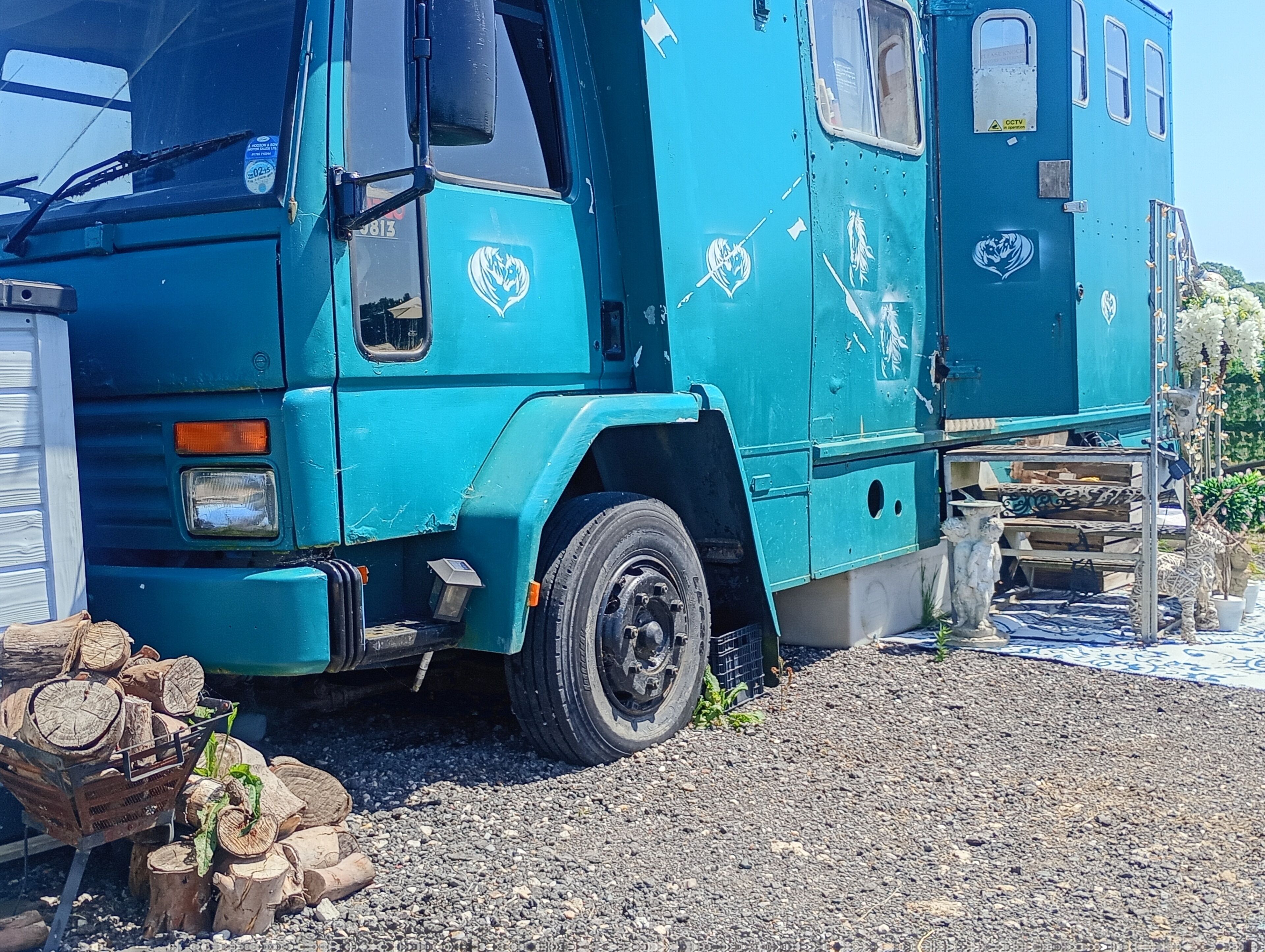 Un camió blau gran amb una cabina, estacionat sobre un sòl de grava, amb troncs i una taula petita a prop.