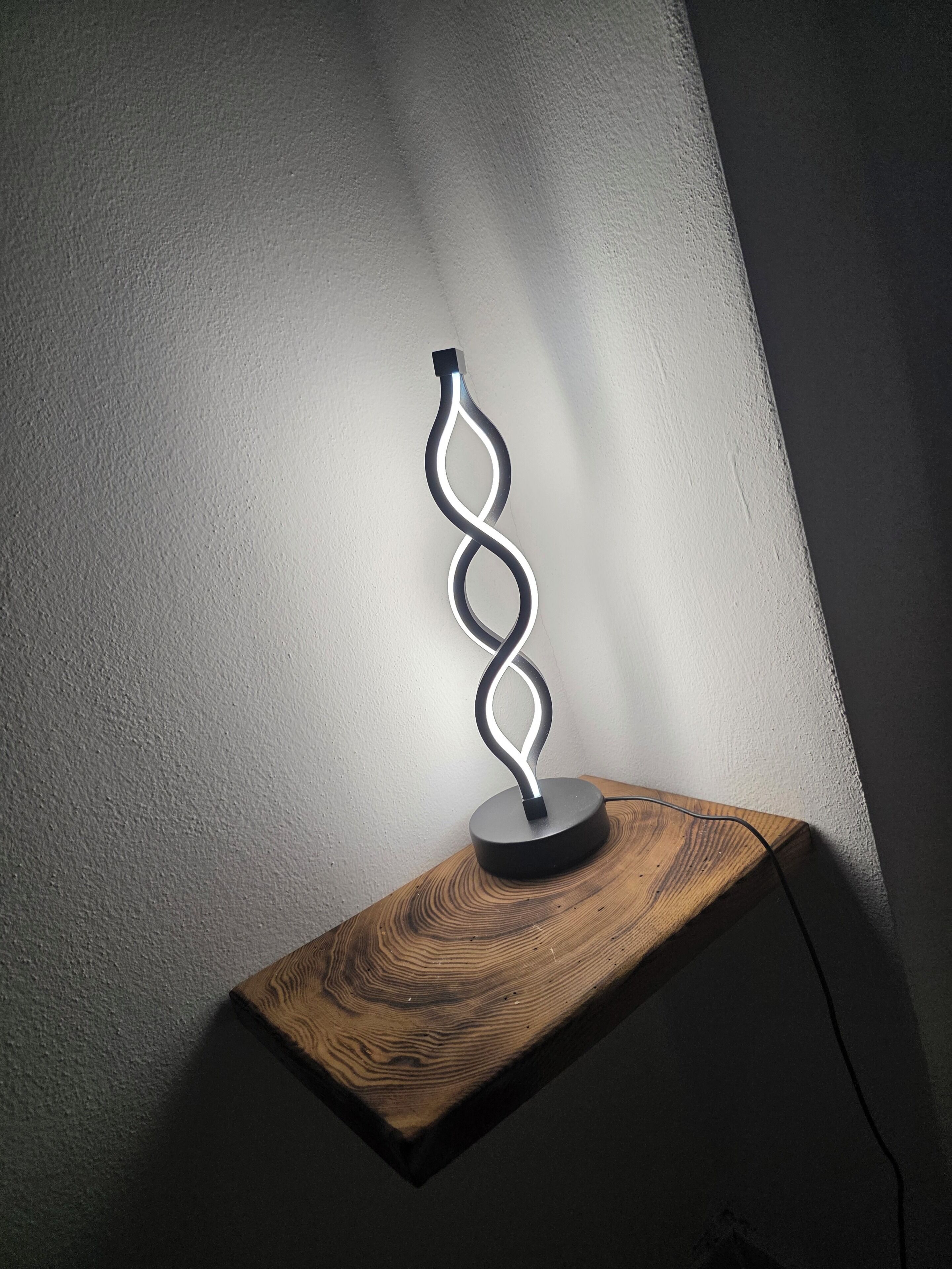 A modern light fixture with a spiral design on a wooden shelf against a plain wall.