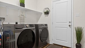Zona de lavandería con dos lavadoras Samsung, una puerta blanca y una pequeña planta en la pared.