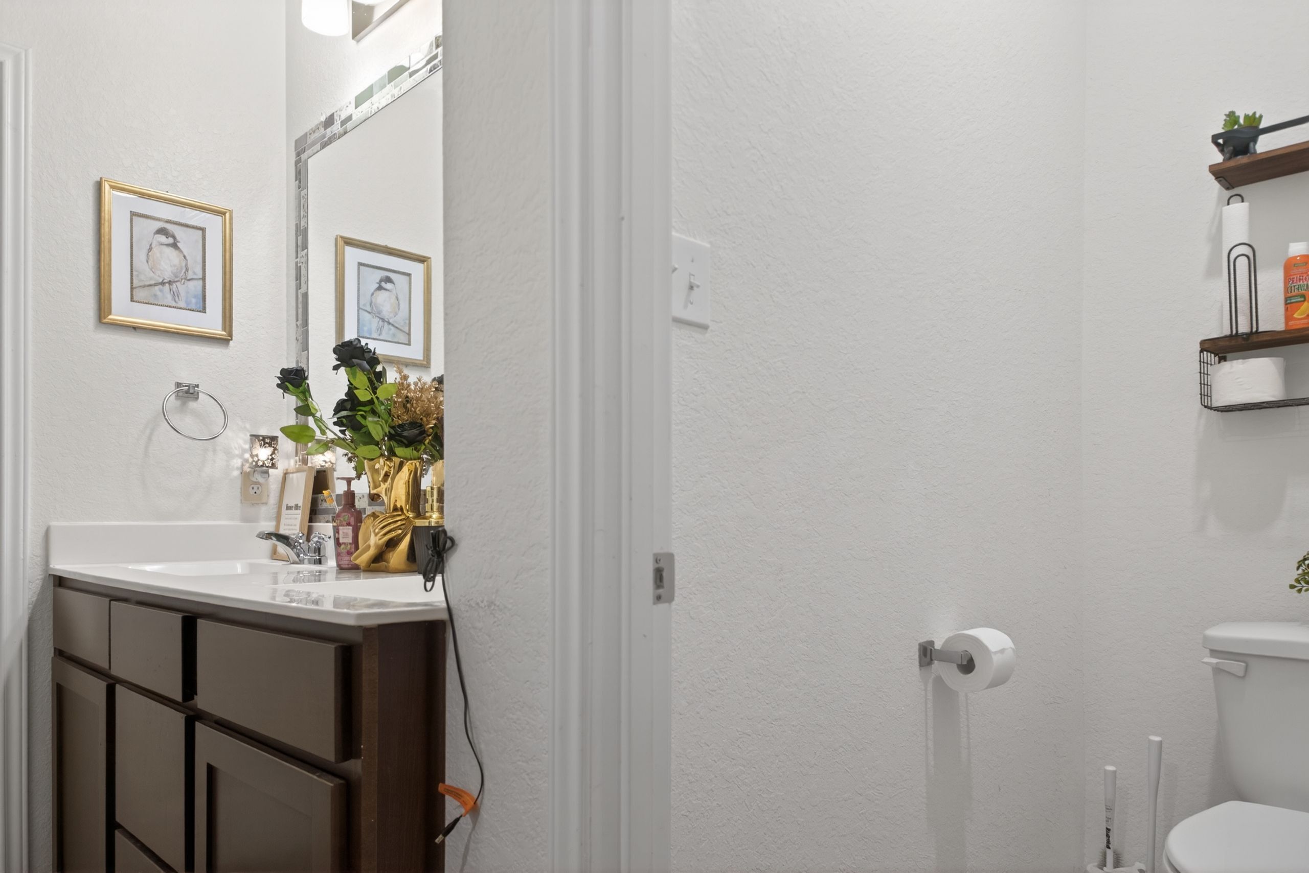 A bathroom with a white sink, dark wooden cabinets, a mirror, framed artwork, a toilet, and a shelf with toiletries.