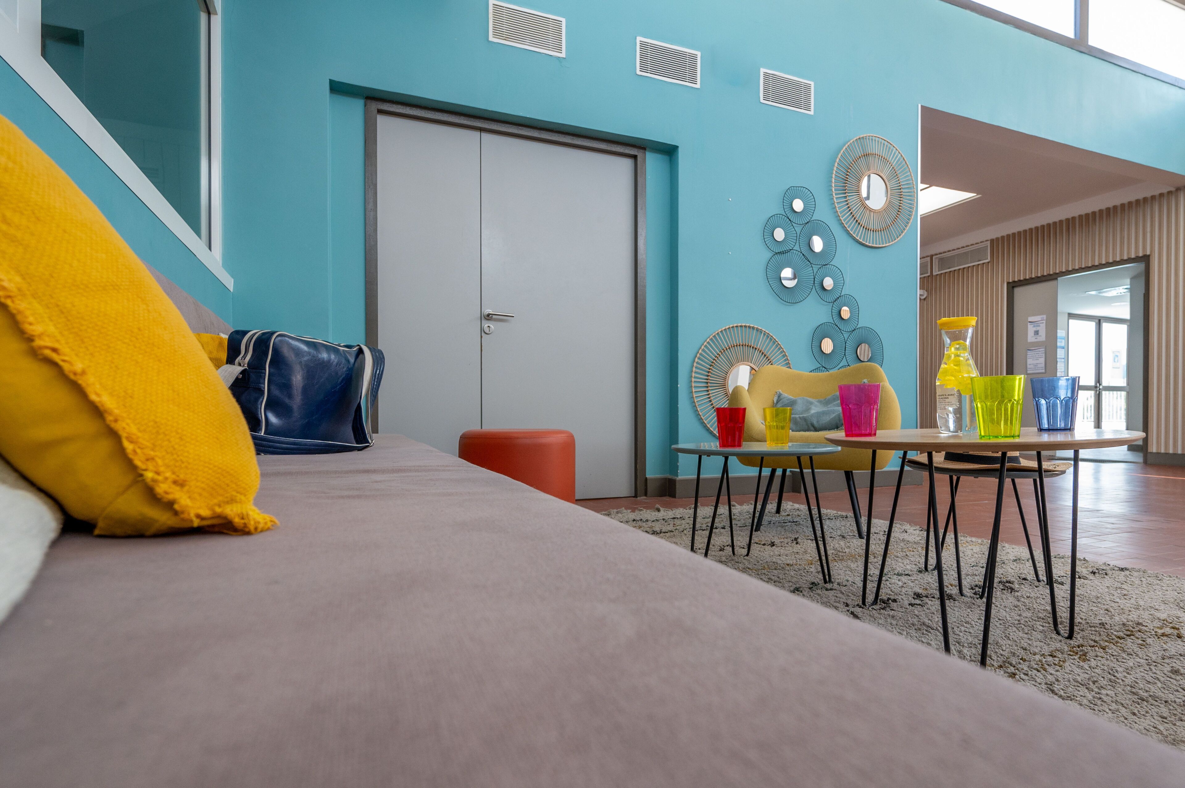 Une pièce avec un lit, une table sur laquelle sont posées des tasses colorées, une chaise et une porte.