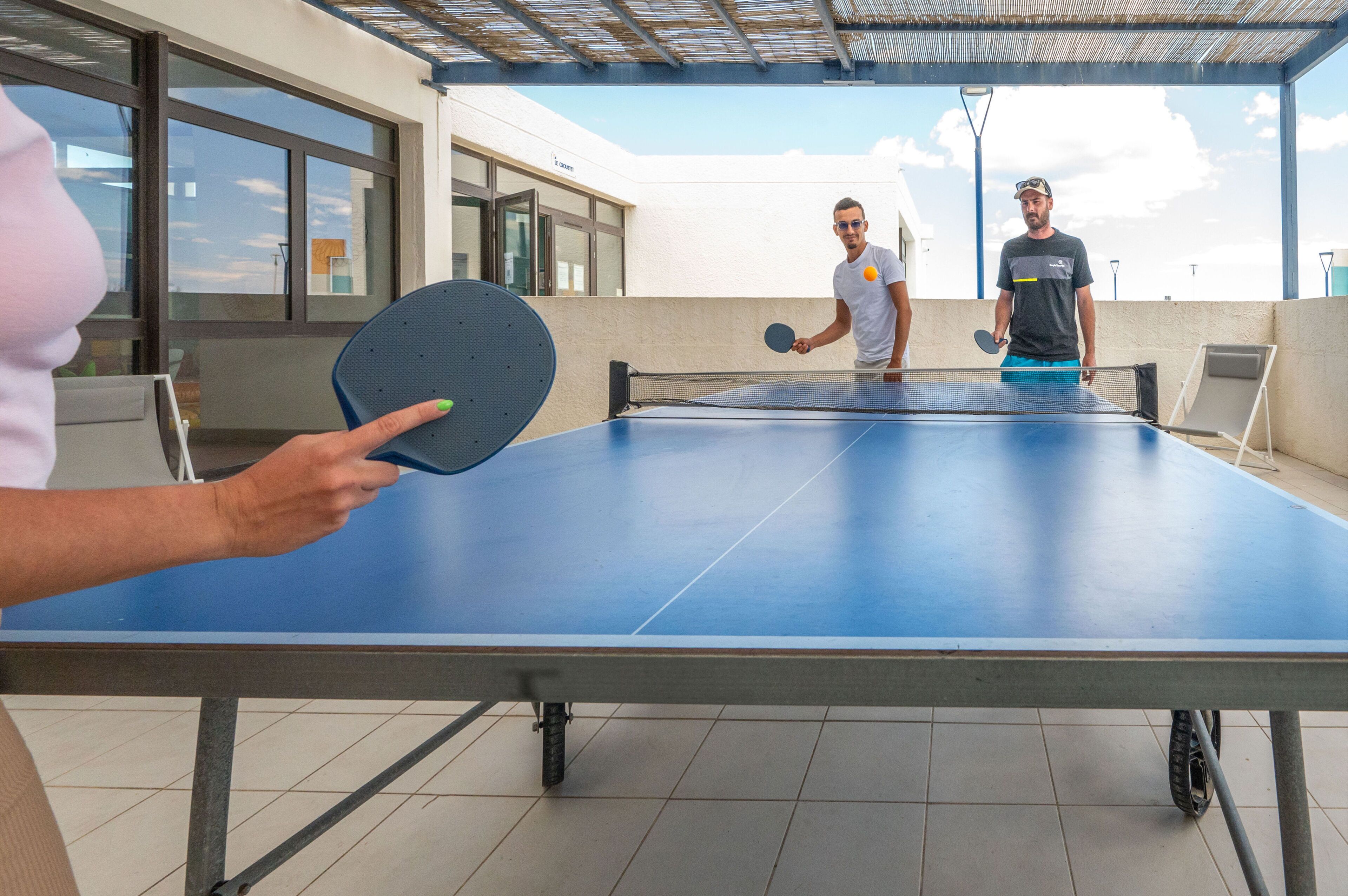 An outdoor ping pong table with two people playing, a building with large windows in the background, and a clear sky.