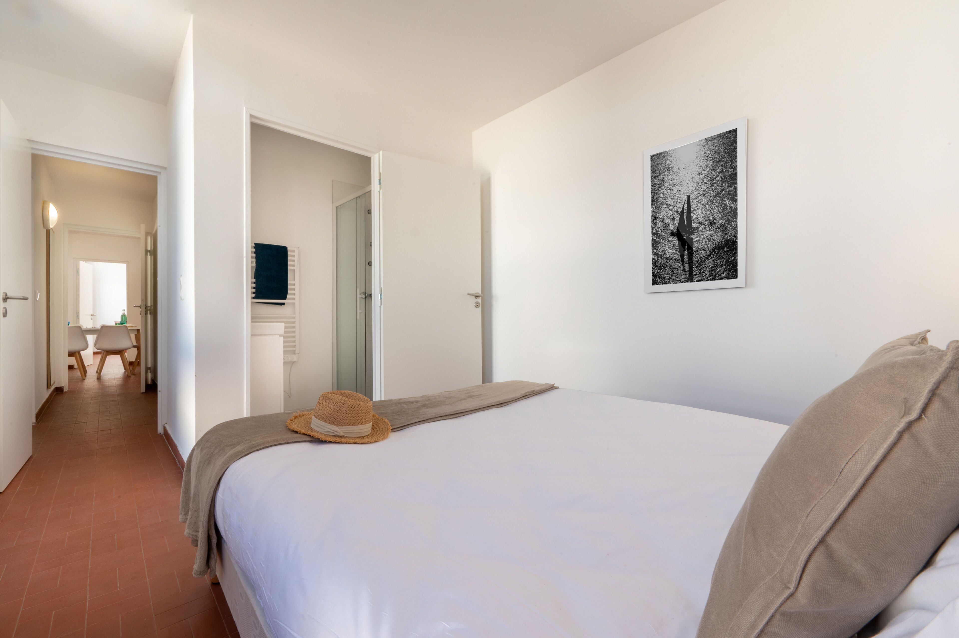Une chambre à coucher avec un lit, un chapeau, un oreiller, un tableau accroché au mur et une porte donnant sur une autre pièce.