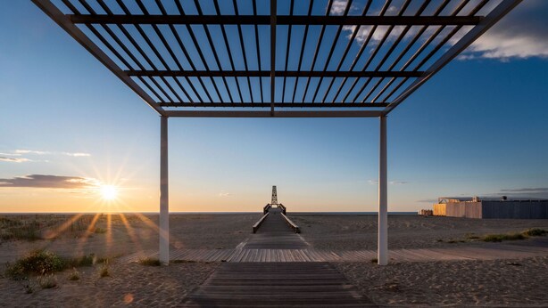 A wooden pier leading to a beach with a metal pergola overhead.