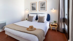 A neatly made bed with a hat, a small bedside table with a book, and two framed pictures on the wall.