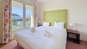 A hotel room with a large bed, two pillows, a green headboard, a nightstand with a lamp, and a view of the outside.