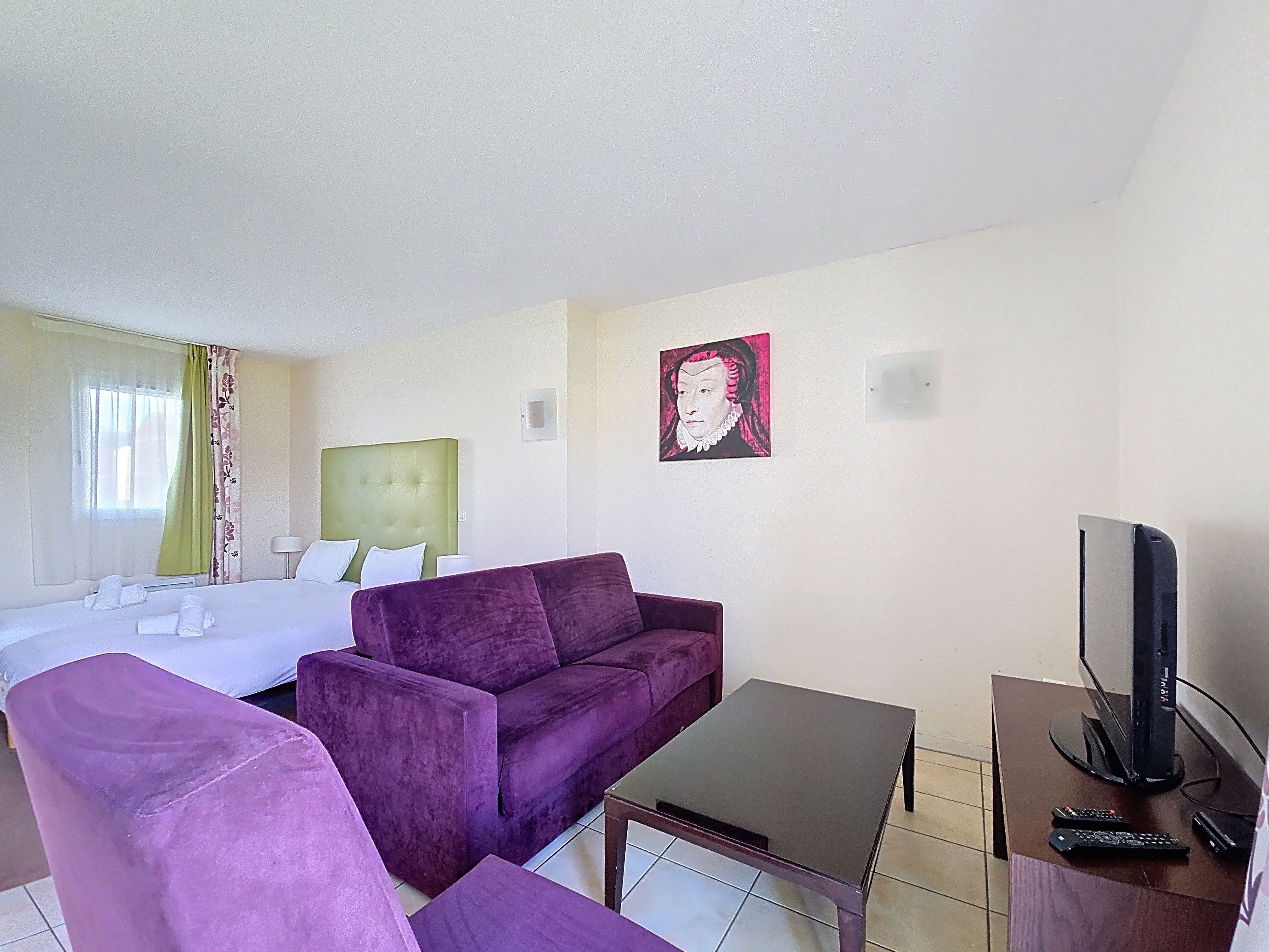 Ein Hotelzimmer mit einem Bett, einem violetten Sofa, einem kleinen Tisch, einem Fernseher und einem Bild an der Wand.