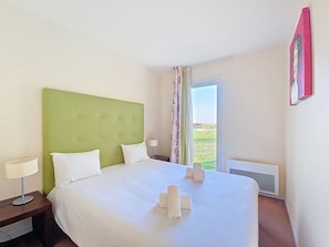 Une chambre d’hôtel avec une tête de lit verte, un lit recouvert de draps blancs, deux lampes de chevet et une vue sur l’extérieur, accessible par une porte-fenêtre coulissante.