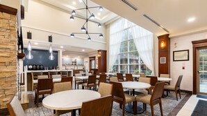 Ein Hotelrestaurant mit runden Tischen und Stühlen, eine Küche im Hintergrund und große Fenster mit Vorhängen.