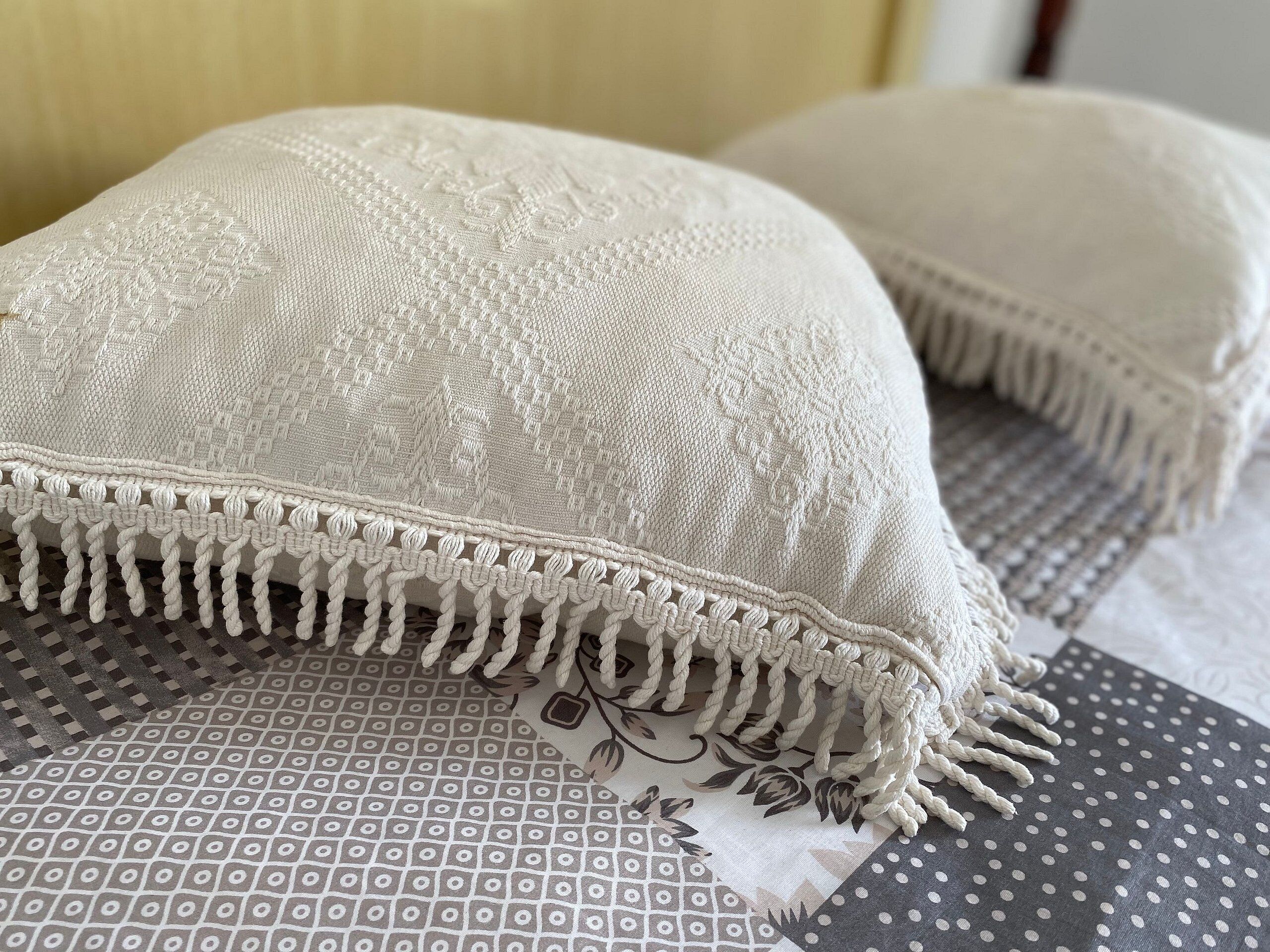 A close-up of a white textured pillow with a fringe detail, placed on a patterned fabric.