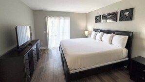 A bedroom with a bed, a dresser, a TV, and a window with blinds. - One Bedroom,  First Floor (Wichita)